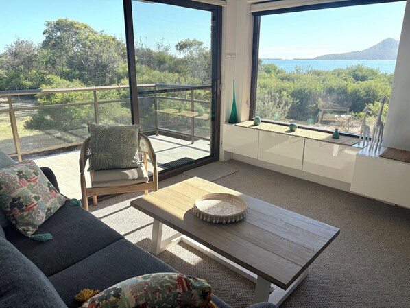 A balcony with a view of the sea and greenery, furnished with a sofa, chair, and table.