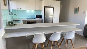 Cozinha moderna com armários brancos, geladeira de aço inoxidável e um balcão longo. Há três cadeiras brancas com pés de madeira.