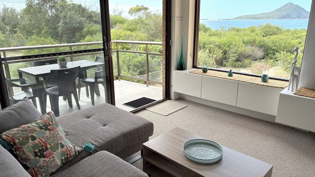 Uma varanda com mesa e cadeiras, um sofá com almofada decorativa, um assento junto à janela com objetos decorativos e vista para área verde e uma montanha ao longe.