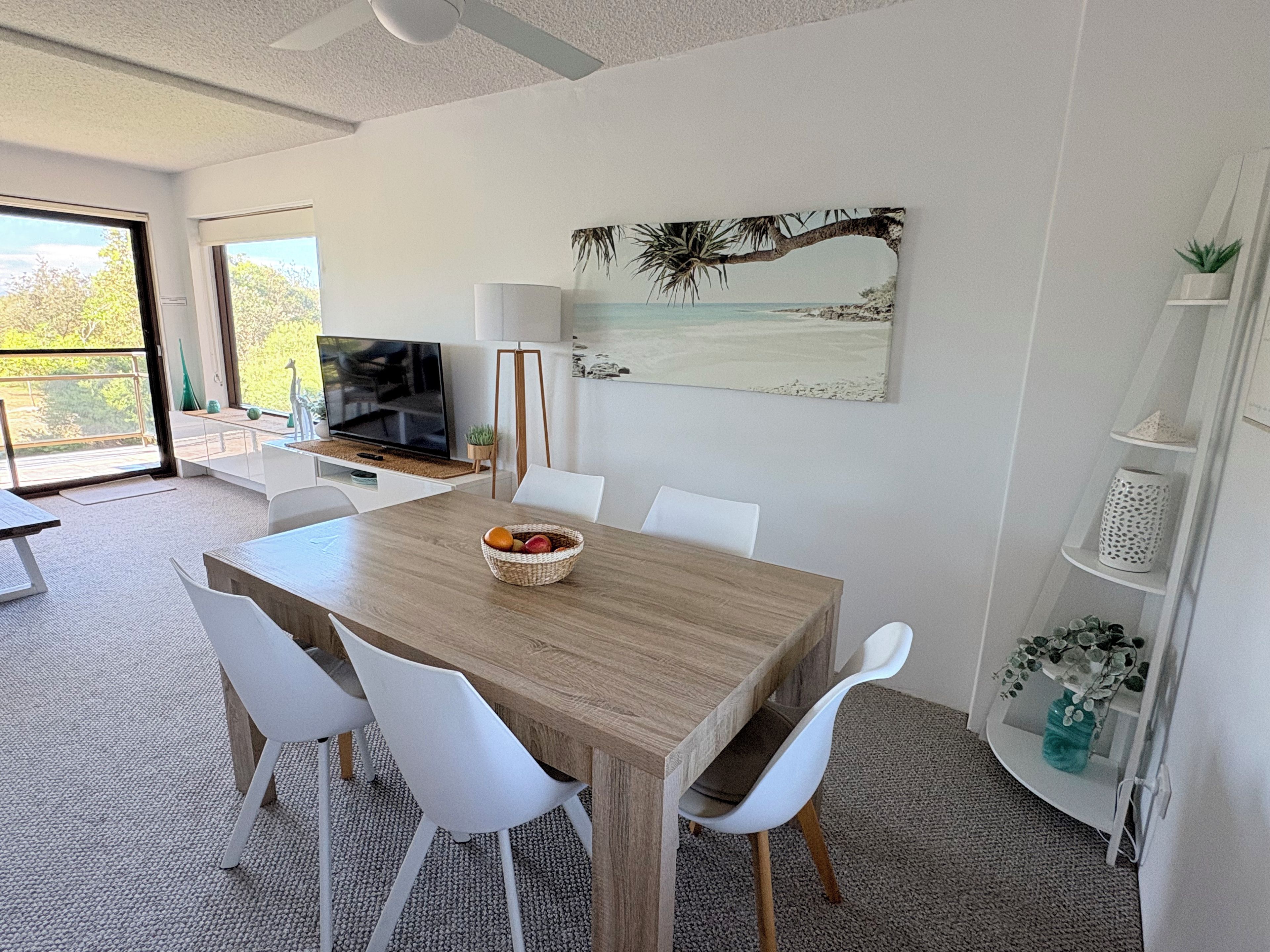 Un comedor moderno con una mesa de madera, sillas blancas, un cuenco de frutas, un televisor y un cuadro con temática marina en la pared.