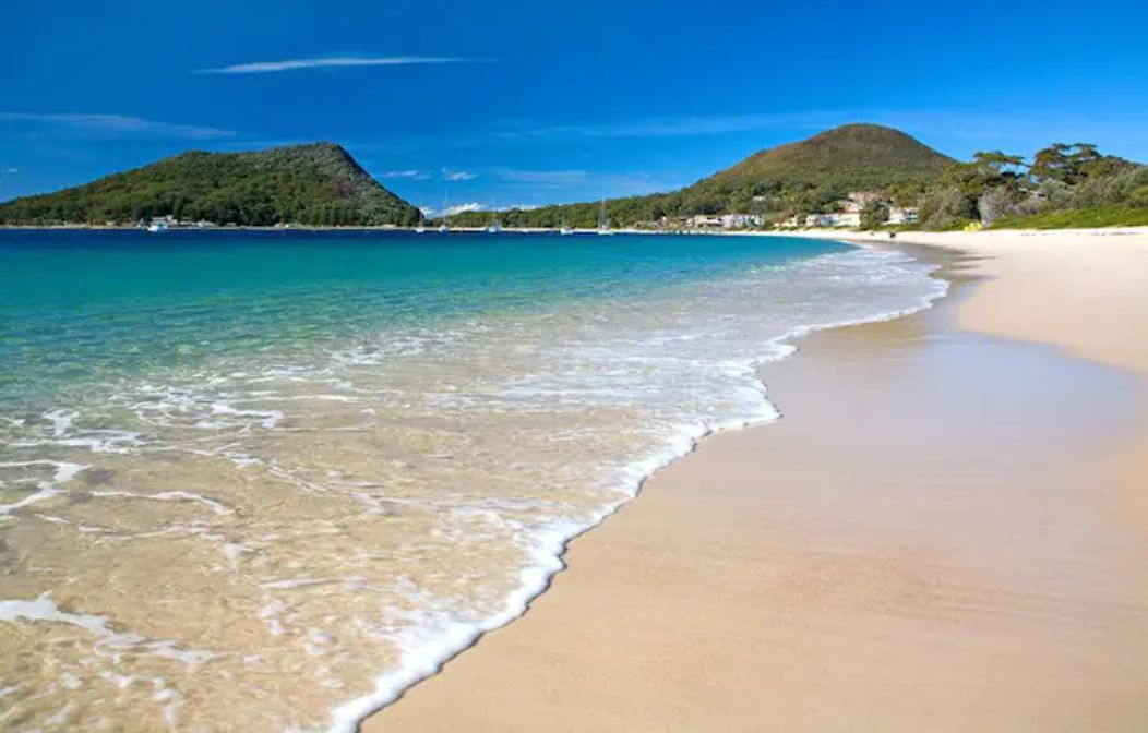 A beach with clear turquoise water, sandy shore, and a hilly landscape in the background.