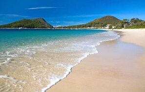 A beach with clear turquoise water, sandy shore, and a hilly landscape in the background.