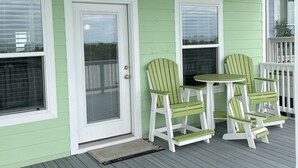 Un porche verde con una mesa y cuatro sillas Adirondack, una puerta y ventanas.