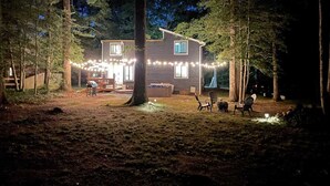 A house with a deck, surrounded by trees, and string lights. - Light & airy home w/private hot tub on golf course (Quinton)