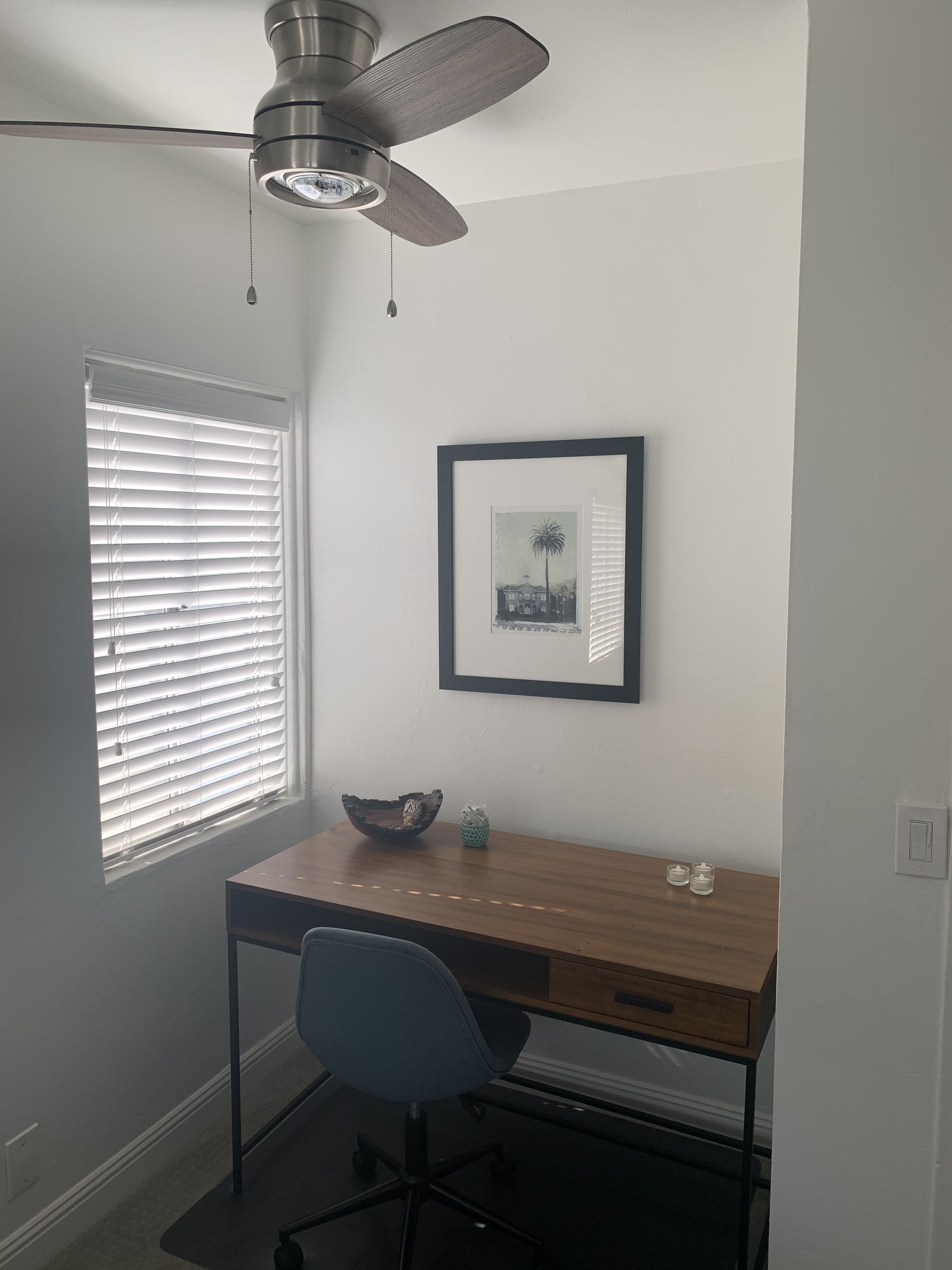 Ein kleiner Büroraum mit Schreibtisch, Stuhl, Deckenventilator, Fenster mit Jalousien, gerahmtem Bild und einem Tisch mit Dekoration.