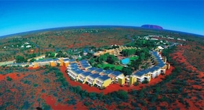 An aerial view of a resort with multiple buildings arranged in a semi-circular layout, surrounded by red vegetation and a clear blue sky.