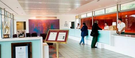 A hotel reception area with a modern design, including a reception desk, framed certificates, and a large abstract painting on the wall.
