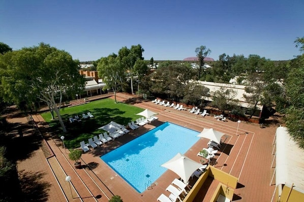 A large outdoor swimming pool with sun loungers and umbrellas, surrounded by trees and a clear sky. - Desert Gardens Hotel (Yulara)