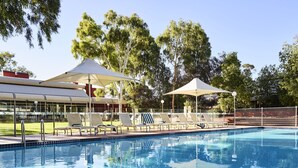 Una piscina con sillas de descanso y sombrillas, rodeada de árboles y un edificio con amplias ventanas.