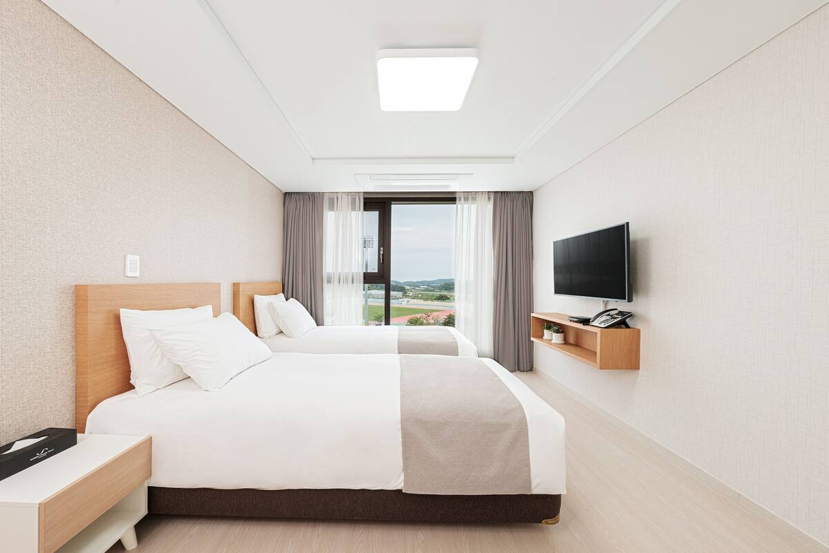 A hotel room with two beds, a TV, a bedside table, a phone, and a view of the outdoors.
