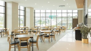Un comedor amplio con mesas y sillas de madera, ventanas grandes que ofrecen vistas de la ciudad, y una cocina moderna visible a través de las ventanas.