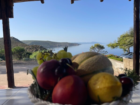 Ein Tisch mit einer Obstschale im Vordergrund und Blick auf eine Küstenlandschaft im Hintergrund.