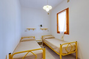 A small, single-room accommodation with two single beds, a bedside table, a shelf with a plant, and a window with curtains.