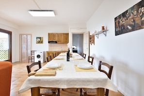 A dining area with a long table set for six, wooden chairs, a kitchen in the background, and a painting on the wall.