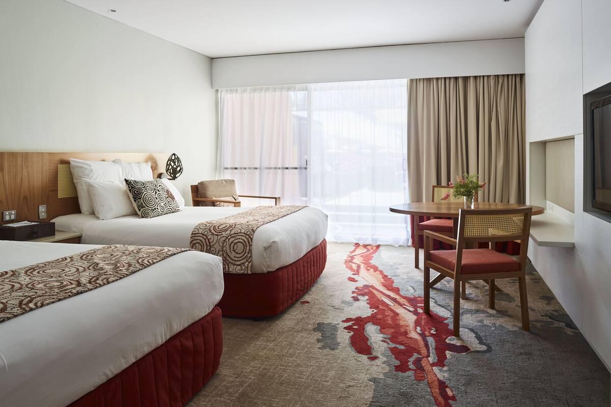 Habitación de hotel con dos camas, una pequeña mesa con sillas, un televisor y un amplio ventanal con cortinas.
