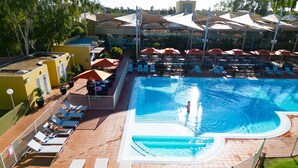 Una amplia piscina con tumbonas y sombrillas, rodeada por una zona vallada con vistas a los edificios circundantes.