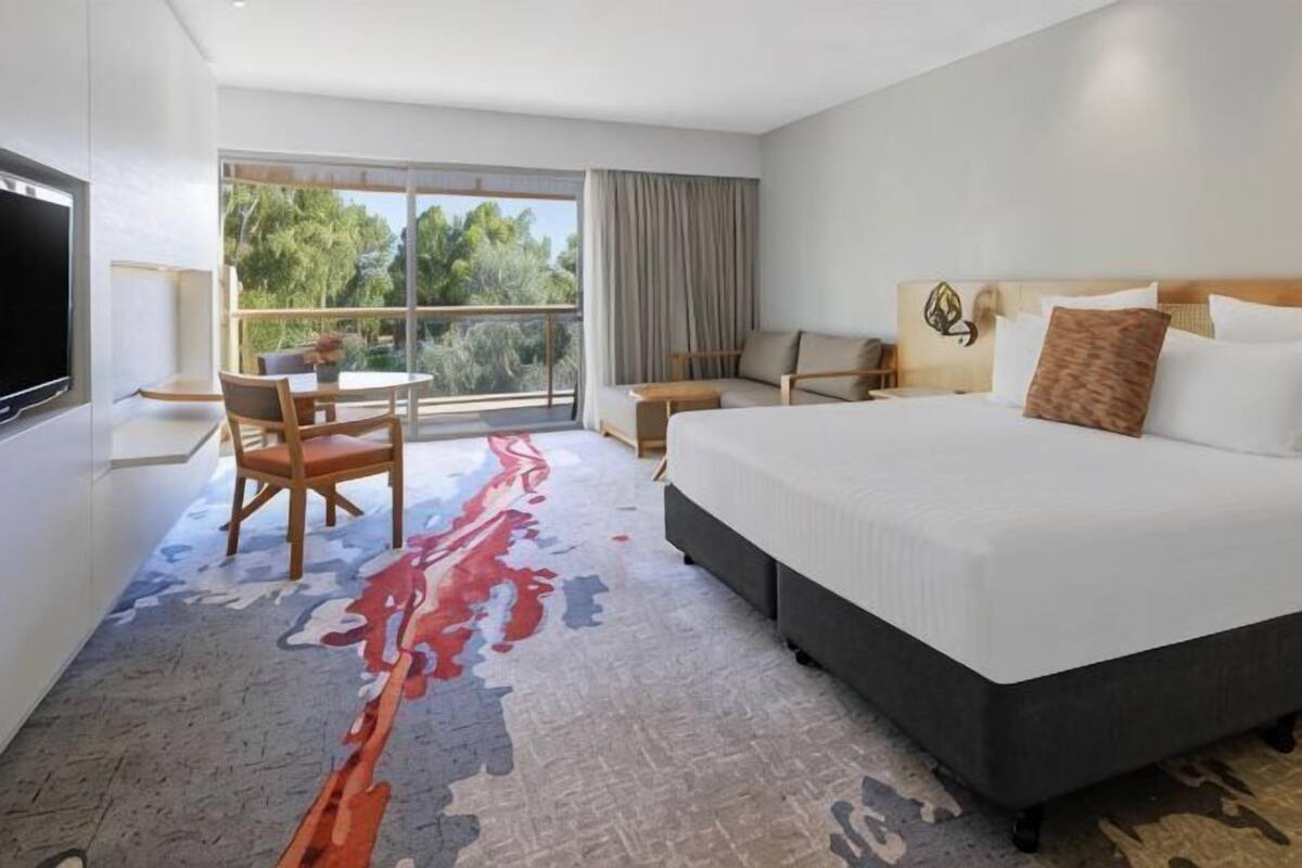 Habitación de hotel con una cama grande, un escritorio, una silla, un televisor y una ventana con vistas a árboles.