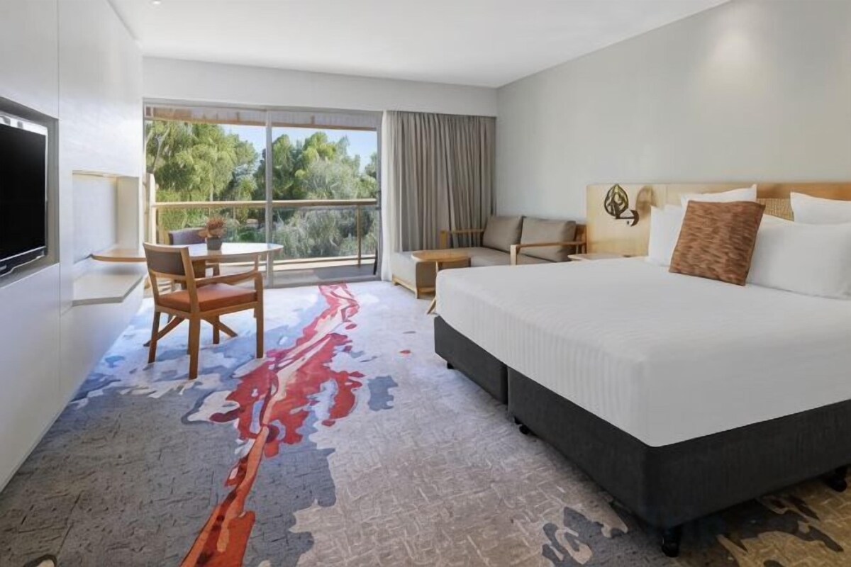 A hotel room with a large bed, a desk, a chair, a TV, and a window with a view of trees.