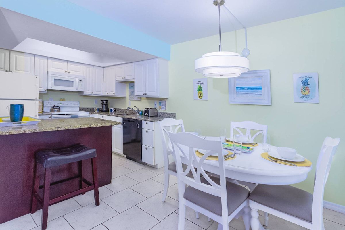 A kitchen with white cabinets, a microwave, a coffee maker, and a dining area with white chairs and a table set for two.