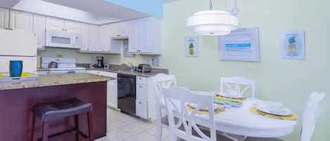 A kitchen with white cabinets, a microwave, a coffee maker, and a dining area with white chairs and a table set for two.