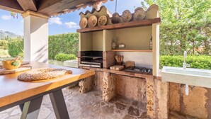 An outdoor kitchen with a grill, a shelf with decorative plates, a wooden table with a woven mat, and a stone floor.