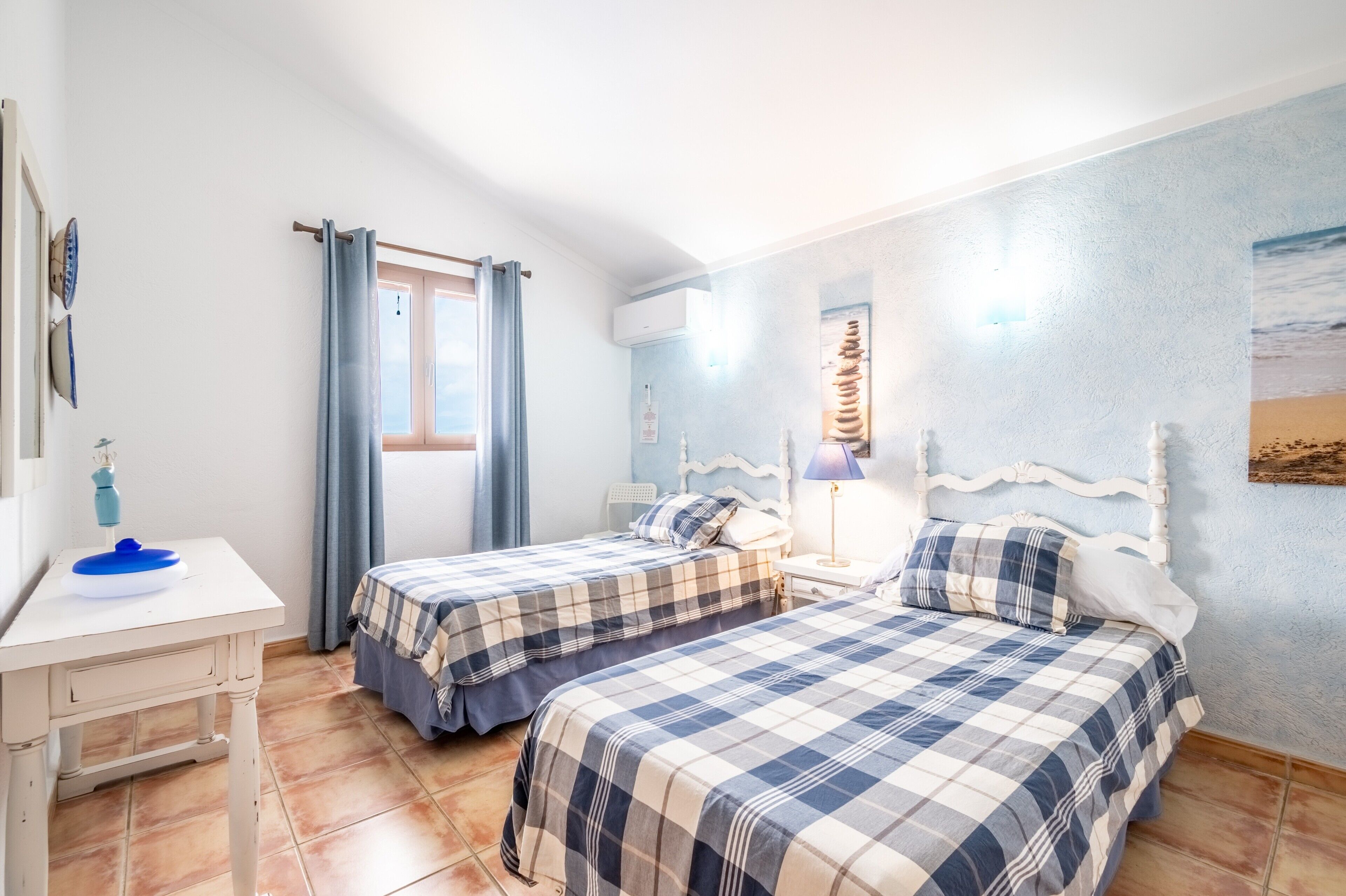 A room with two beds, a small table, a window with curtains, and a wall with a painting.