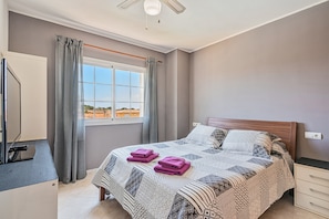 Un dormitorio con cama, ventana con cortinas, mesita de noche y televisor montado en la pared.