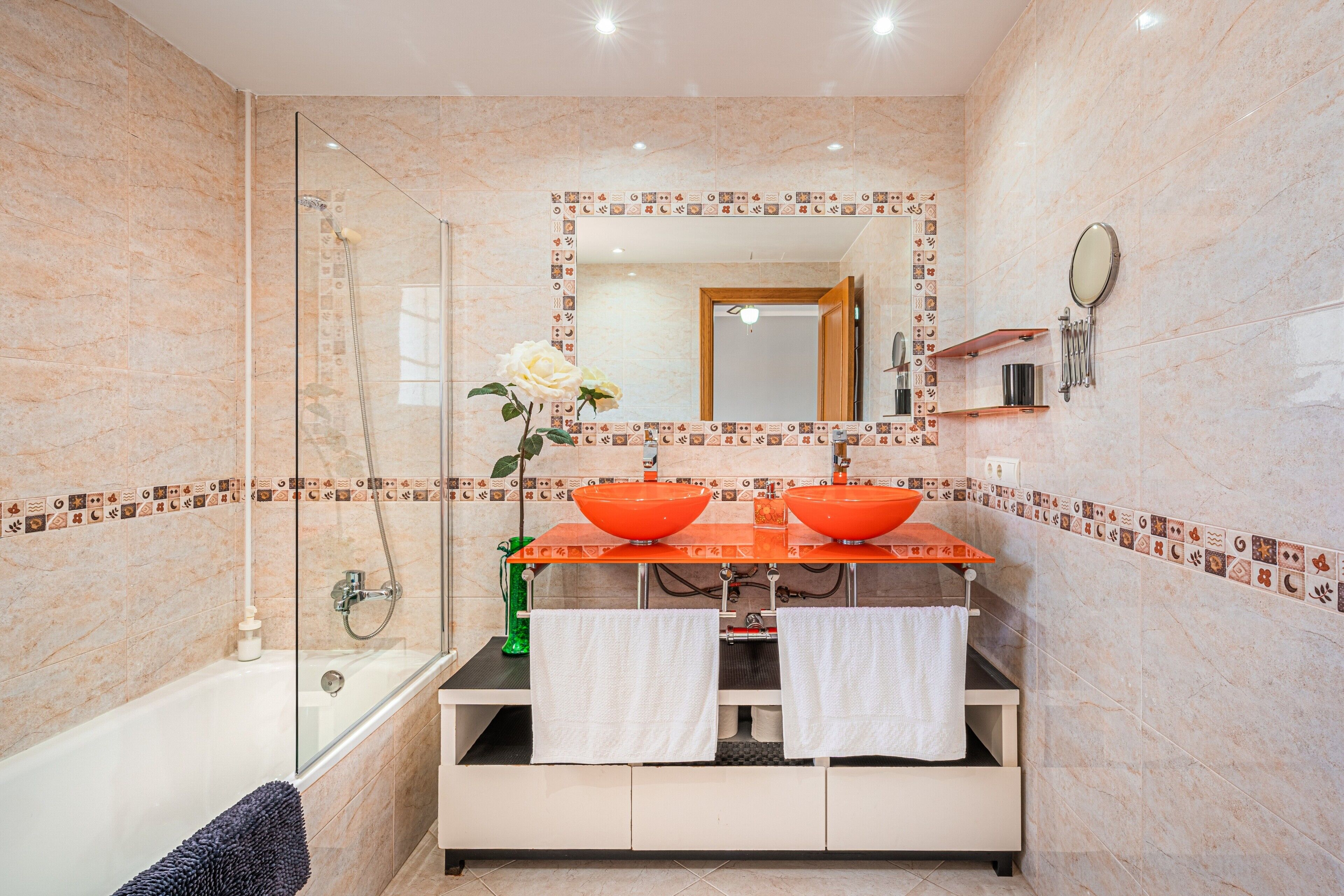 Een badkamer met een douche met glazen wand, twee wastafels met oranje kommen, een spiegel en een decoratieve tegelrand.