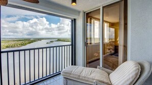 A balcony with a view of a river and a cityscape, a wicker chair, and a sliding glass door leading to an interior space. - Ocean Harbor #1201B Beautiful Bayfront Condo (Fort Myers Beach)