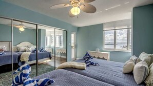 A bedroom with two beds, a ceiling fan, a large window with a view, and a glass partition. - Ocean Harbor #1201B Beautiful Bayfront Condo (Fort Myers Beach)