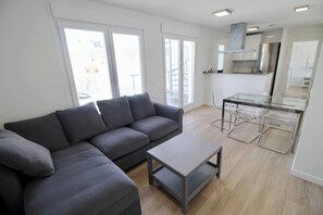 A modern living room with a grey sofa, a glass coffee table, and a kitchen area with a stainless steel refrigerator and a glass dining table.