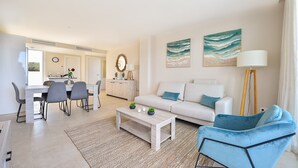 A modern living room with a sofa, coffee table, and chairs. There is a dining area with a table and chairs. The room has artwork on the walls and a floor lamp.