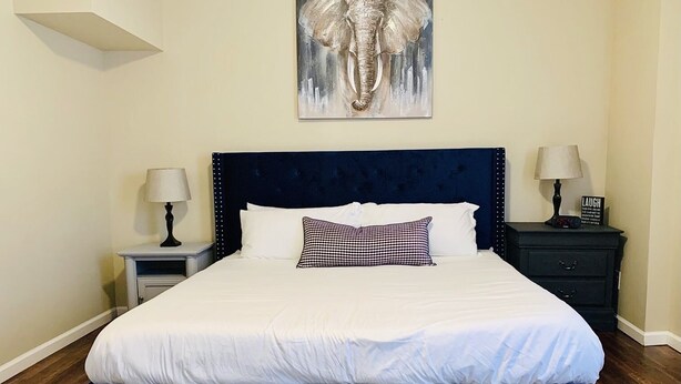 A bedroom with a large bed, two bedside tables, a nightstand, a wall-mounted artwork of an elephant, and a dark blue headboard.