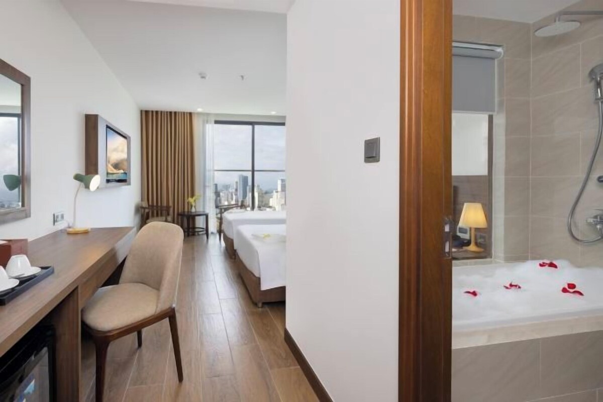 A hotel room with a wooden desk, a chair, a bed, a bathroom with a bathtub, and a city view.