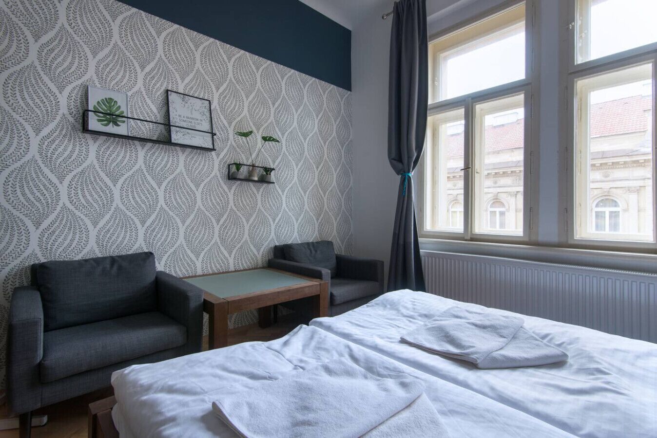 A hotel room with a bed, two armchairs, a small table, a window with curtains, and a patterned wallpaper.