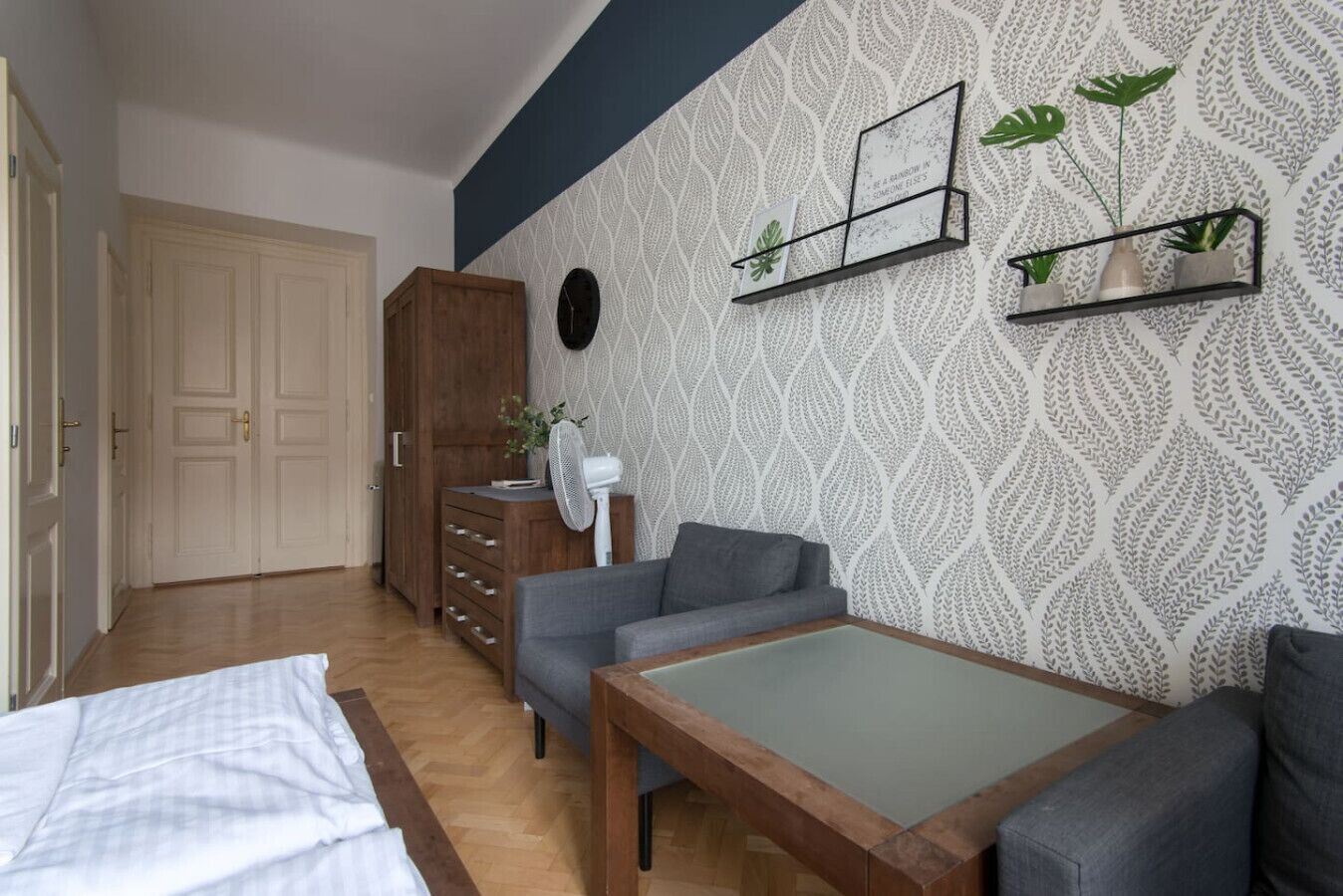 Une pièce comprenant un lit, un canapé gris, une table en bois, une armoire et un mur orné d’un motif de feuilles.