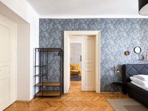 Una habitación con cama, un estante metálico negro, una puerta que da a otra estancia y una pared con textura.