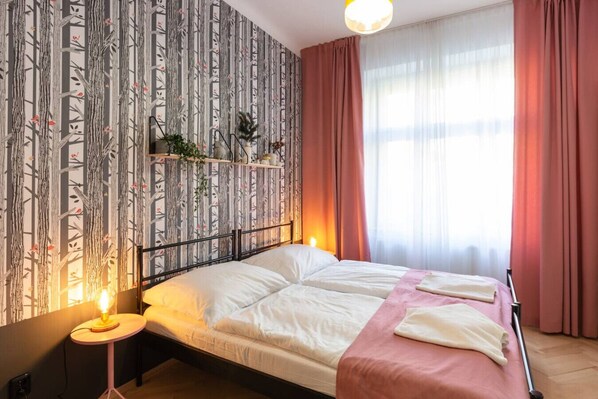 Une chambre à coucher avec un lit, une table de chevet avec une lampe, une fenêtre avec des rideaux et un mur recouvert d’un papier peint à motifs.