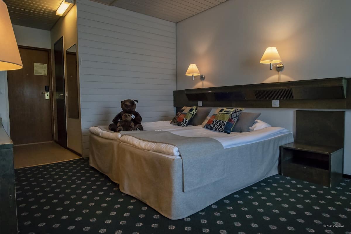 A hotel room with a bed, two wall-mounted lamps, a teddy bear, and a small table.