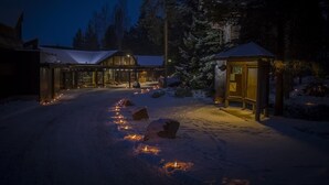 Un sendero cubierto de nieve que conduce a una cabaña con fachada de vidrio y una pequeña estructura de madera con techo.