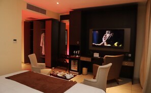 A hotel room with a bed, two armchairs, a TV, a minibar, and a wardrobe.