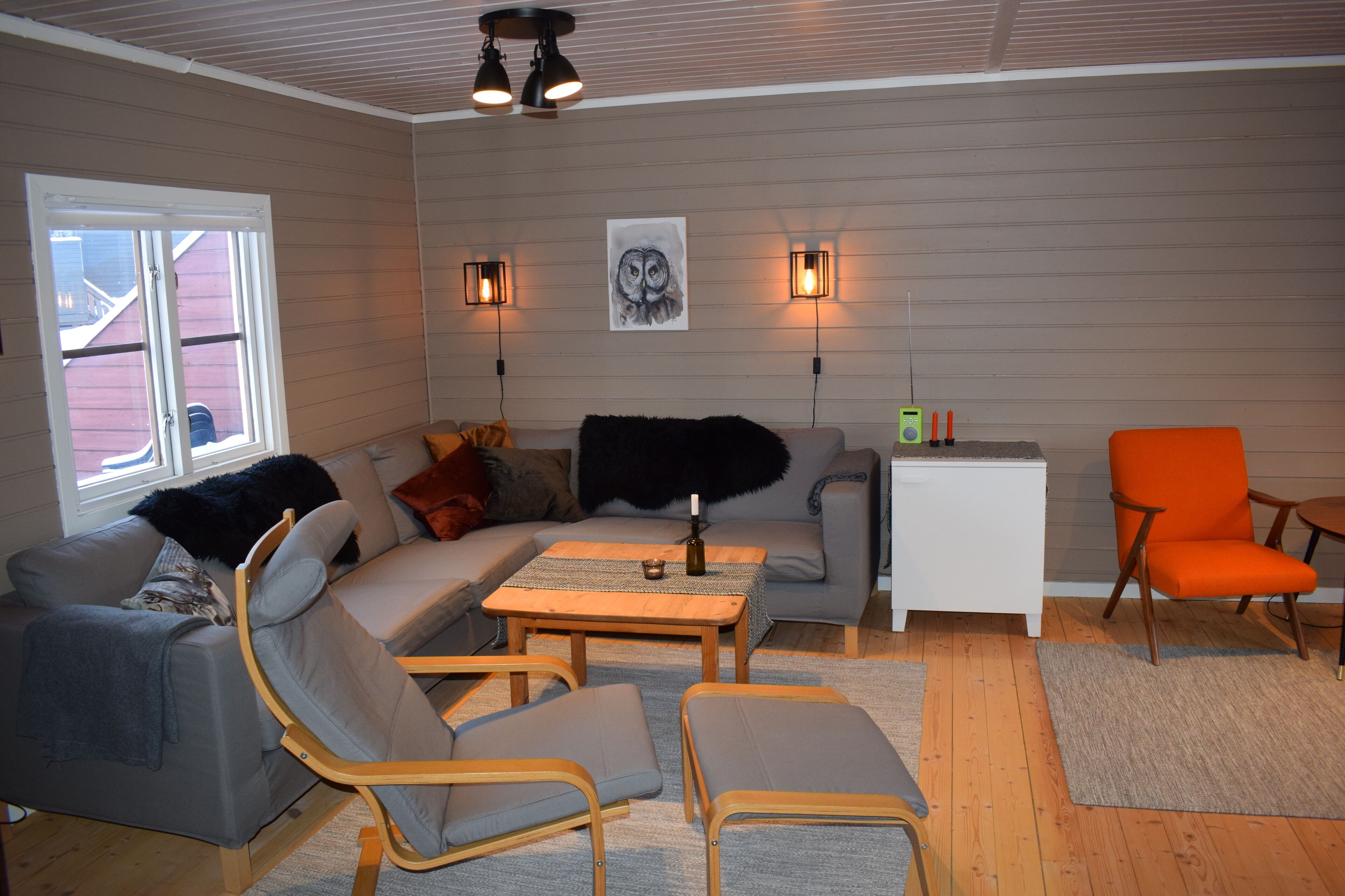En stue med en grå sofa, to lænestole, et sofabord og et lille, hvidt sidebord. Der er to vægmonterede lamper og et billede af en ugle i ramme.