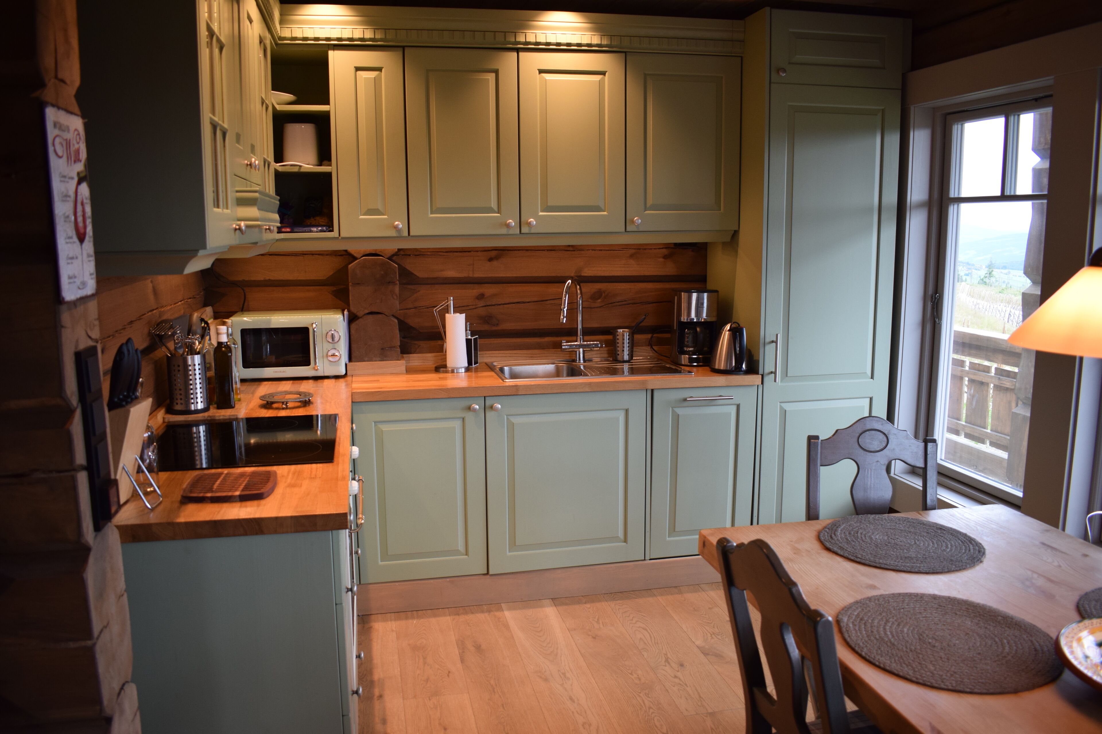 A kitchen with light green cabinets, a wooden countertop, a microwave, a coffee maker, and a window with a view.