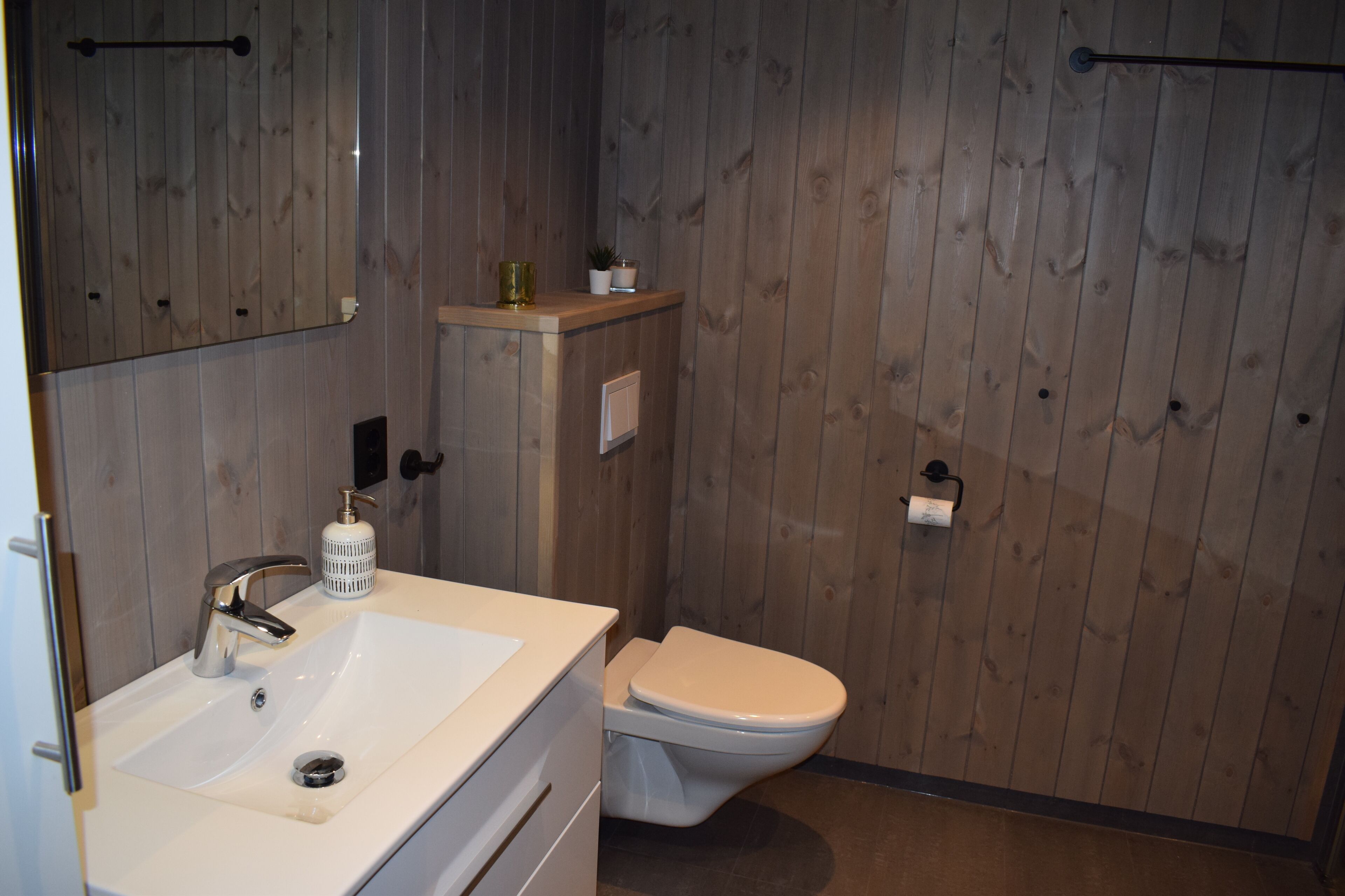 Een badkamer met een houten wand, een wit wastafelmeubel met een chromen kraan, een toilet en een klein plankje met een plant en een fles.