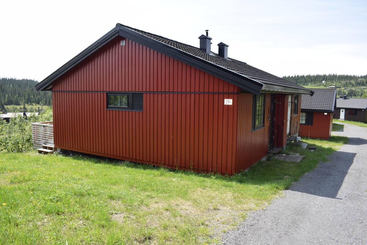 A red wooden cabin with a sloped roof, a small porch, and a gravel driveway.