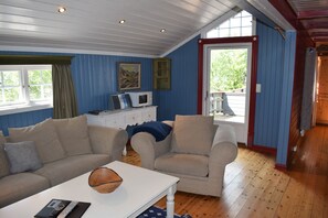 A room with a blue wall, wooden floor, a white table, two beige armchairs, a white dresser, a painting, and a window with curtains. - Skeikampen Booking (Gausdal)