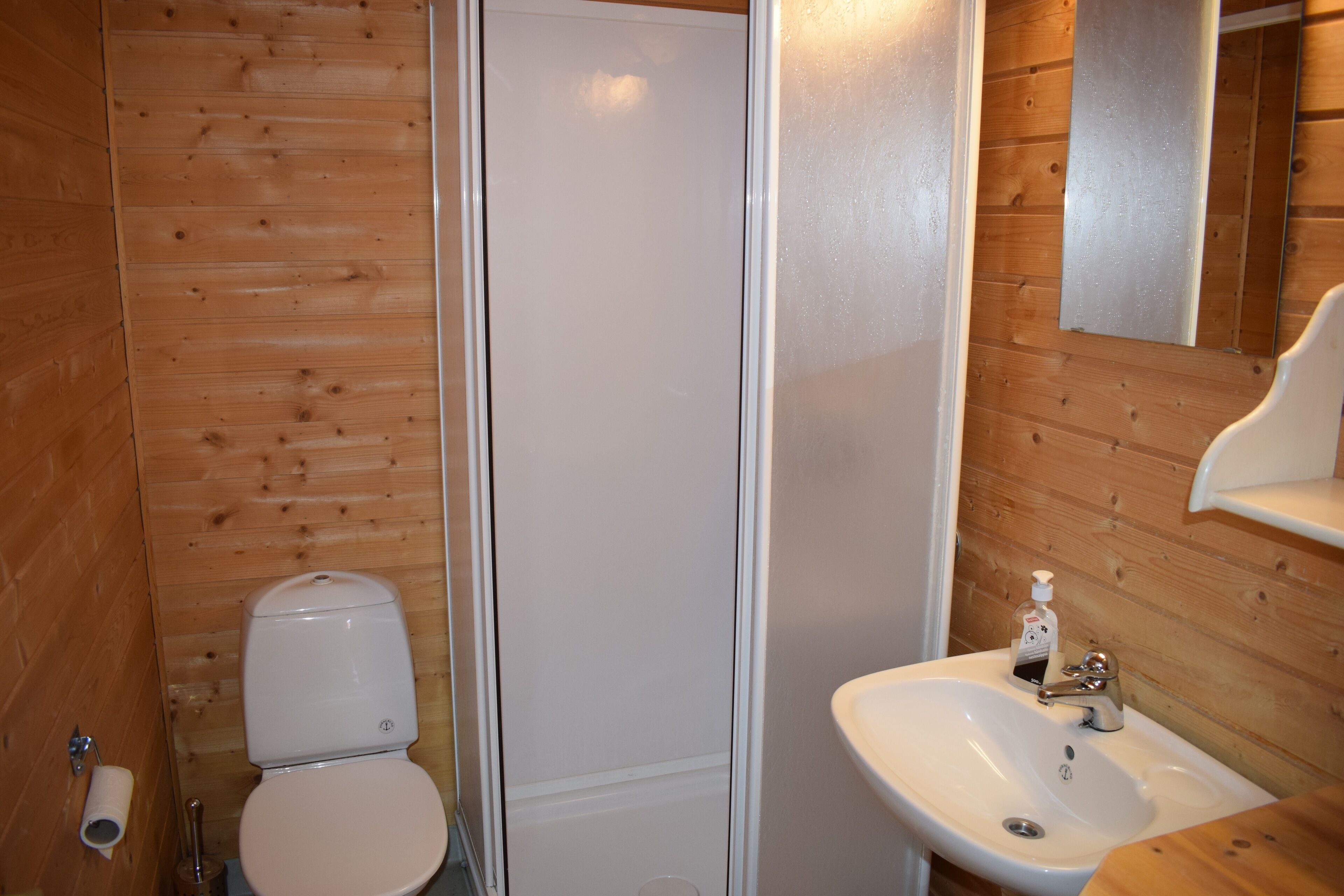 A bathroom with wooden walls, a toilet, a shower, and a sink with a mirror.