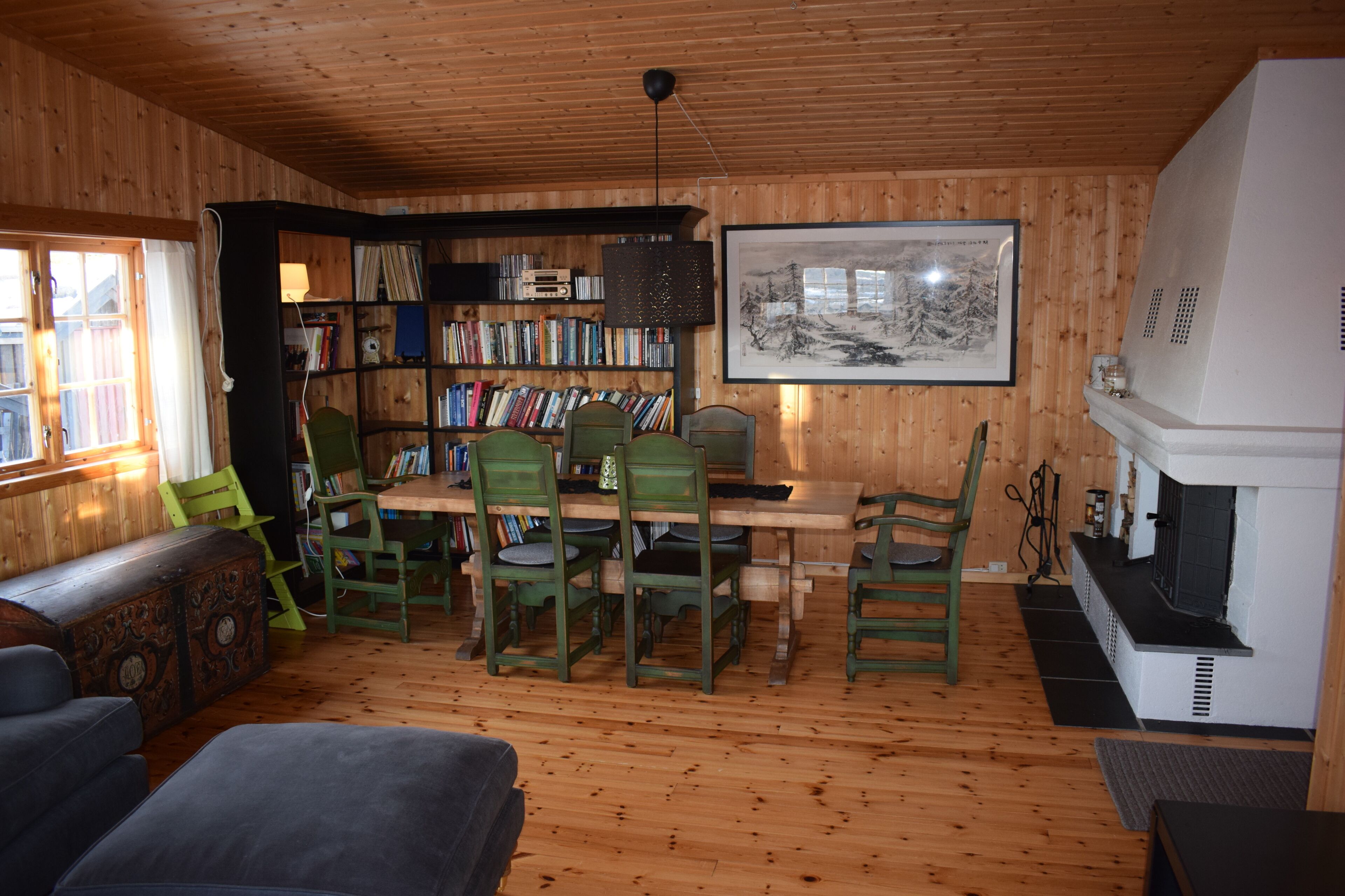 Una habitación acogedora con paredes y suelos de madera, que incluye una mesa de comedor con sillas, una estantería, una chimenea y un ventanal con cortinas.