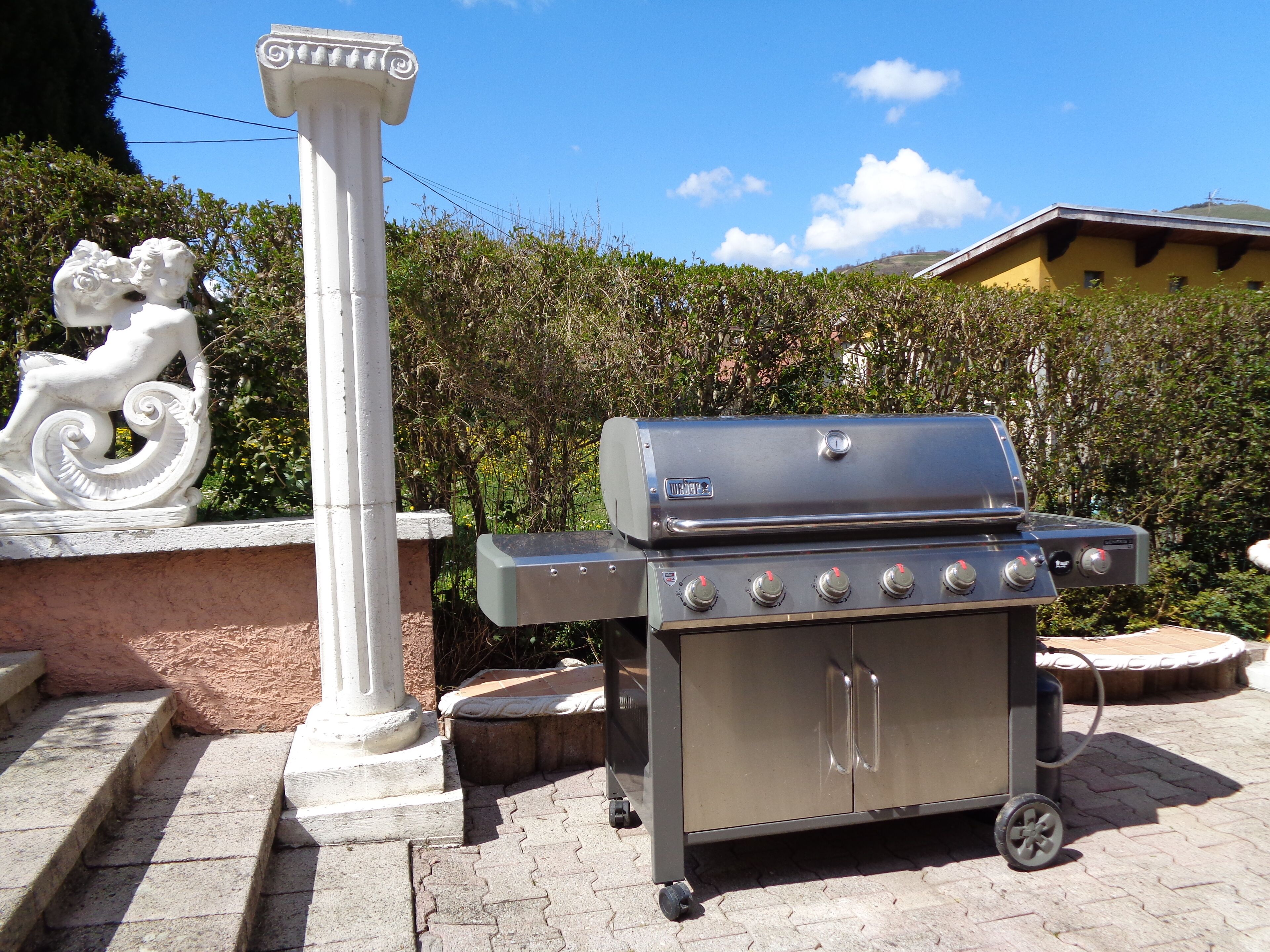 Un barbecue en acier inoxydable doté de quatre brûleurs et d’un meuble latéral, installé sur une terrasse pavée avec des marches menant à une colonne blanche ornée d’une sculpture décorative.