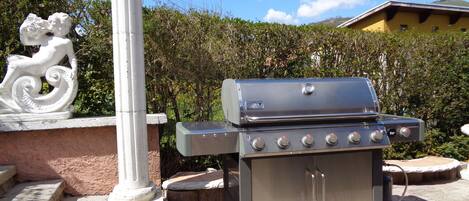 Un barbecue en acier inoxydable doté de quatre brûleurs et d’un meuble latéral, installé sur une terrasse pavée avec des marches menant à une colonne blanche ornée d’une sculpture décorative.